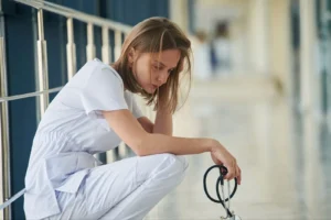 A doctor sitting down, looking stressed, while holding a stethoscope.