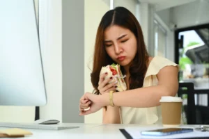 A woman sitting, eating her sandwich, and looking at her watch.