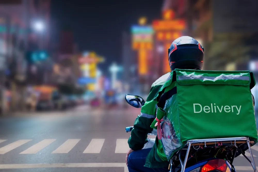 A delivery driver on a scooter with an app-branded backpack riding through a city street.