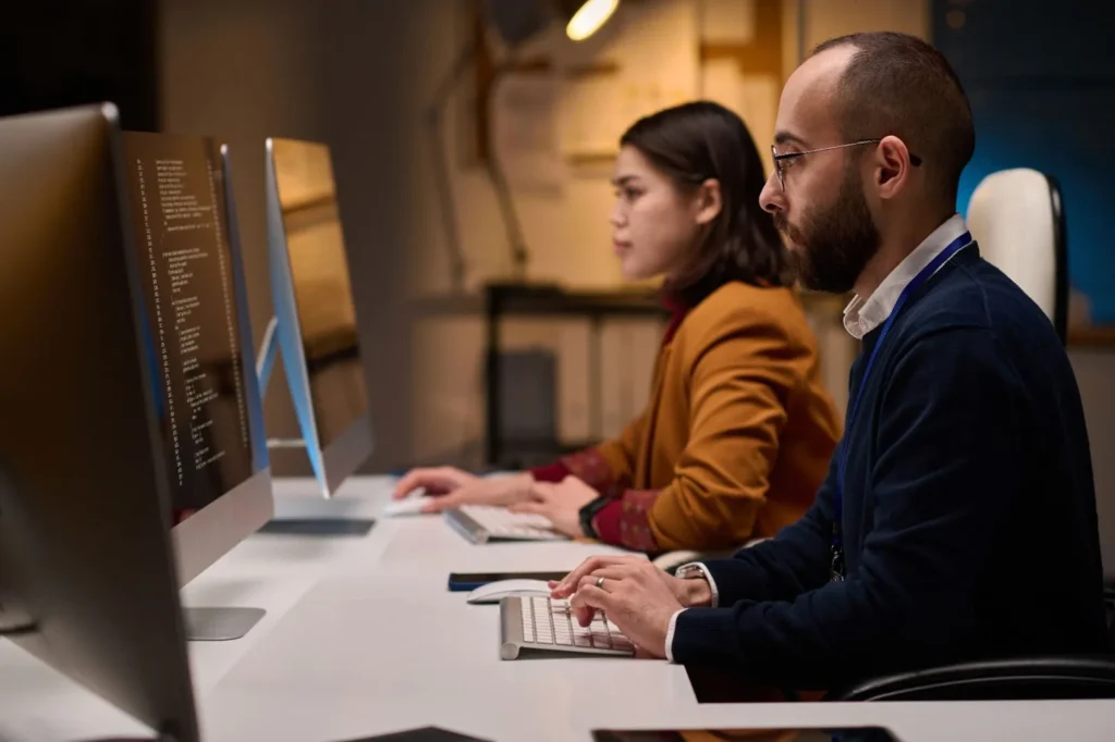 A man and a woman are sitting in front of desktops.